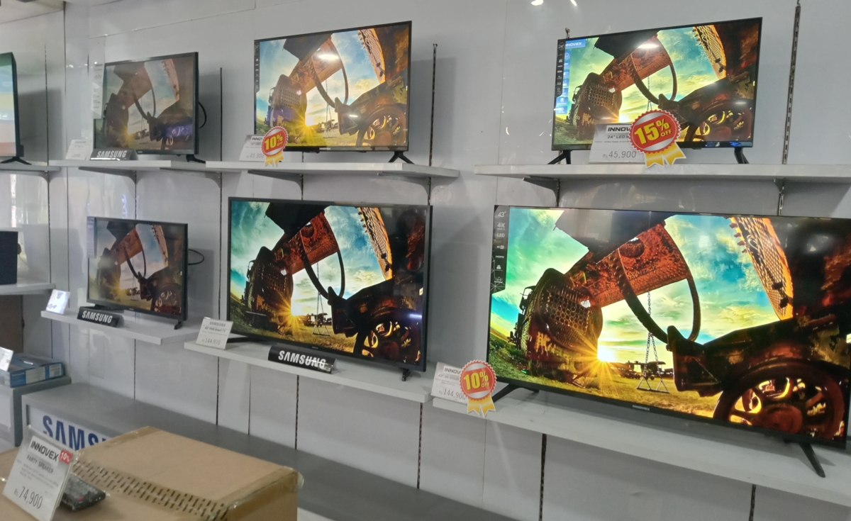 A wide shot of a retail electronics store aisle, showcasing a display of multiple Samsung smart televisions of various sizes mounted on white shelves. All the TVs are displaying the same vibrant, colorful image of what appears to be an ancient, weathered structure or ruin against a bright sky. Some TVs have small orange or red circular price tags with percentages like "10%" or "15%" discounts visible. The "SAMSUNG" brand name is visible on the bottom frame of some of the televisions. In the foreground on the left, part of a cardboard box and another price tag are visible.
