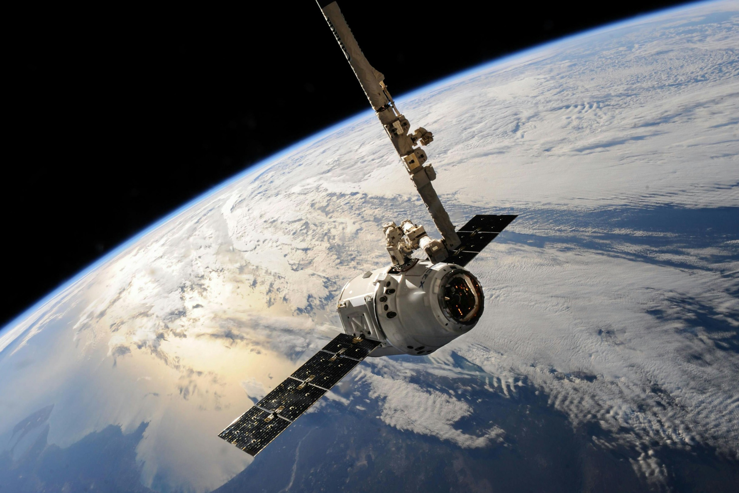 A wide shot of the SpaceX Dragon cargo spacecraft being maneuvered by the International Space Station's Canadarm2 robotic arm. An astronaut in a white spacesuit is visible working on the arm as it holds the capsule high above the Earth's curved horizon, which shows blue oceans and white clouds against the blackness of space.