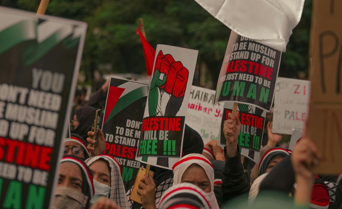 A protest is shown from a low angle, with a large crowd of people holding up signs. The signs have various messages, many of which are related to Palestine. One sign in the center features a Palestinian flag in the shape of a raised fist, with text that reads "DON'T LET PALESTINE TO BE MY WILL BE FREE!!!" Another sign on the right reads "TO BE MUSLIM STAND UP FOR PALESTINE YOU JUST NEED TO BE HUMAN". The crowd consists of people wearing keffiyeh and other head coverings. The overall atmosphere is one of activism and solidarity.