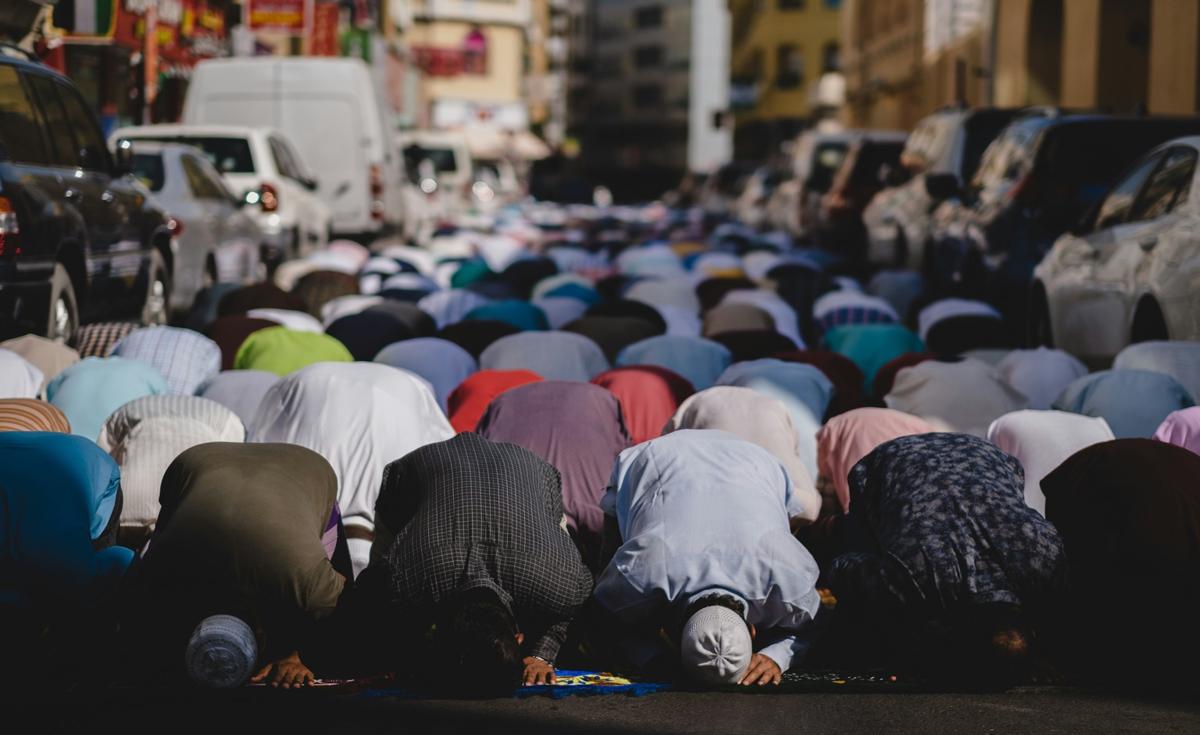 A large group of Muslims wearing colorful clothing is praying together in rows on a city street, with cars parked along both sides.